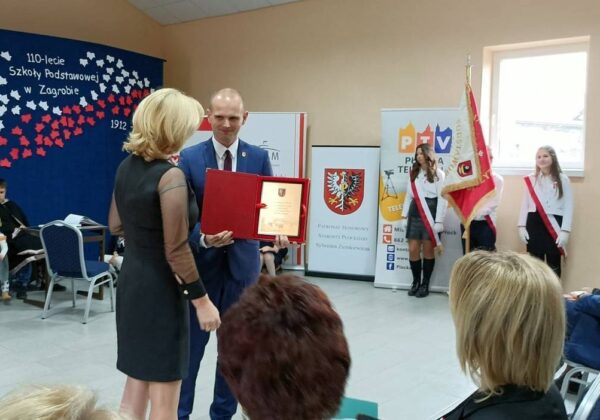 Uroczystość wręczenia wyróżnienia z okazji 110-lecia Szkoły Podstawowej w Zagrobiu, gdzie osoba w eleganckim stroju otrzymuje dyplom od przedstawiciela szkoły.
