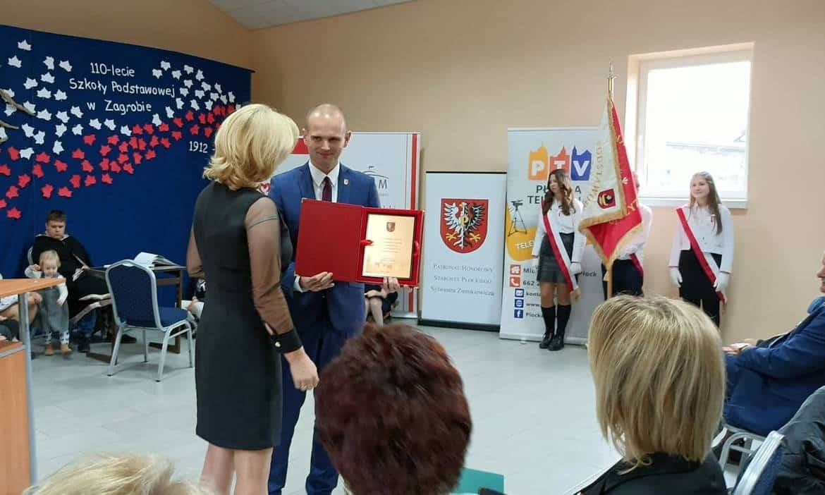 Uroczystość wręczenia wyróżnienia z okazji 110-lecia Szkoły Podstawowej w Zagrobiu, gdzie osoba w eleganckim stroju otrzymuje dyplom od przedstawiciela szkoły.