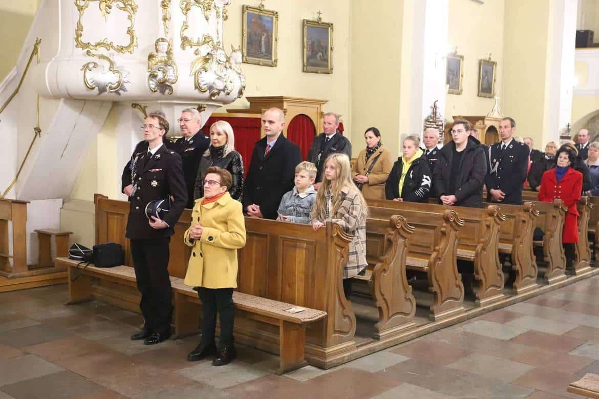 Grupa ludzi w strojach formalnych, w tym dzieci, stoi w kościele, uczestnicząc w ceremonii lub modlitwie.