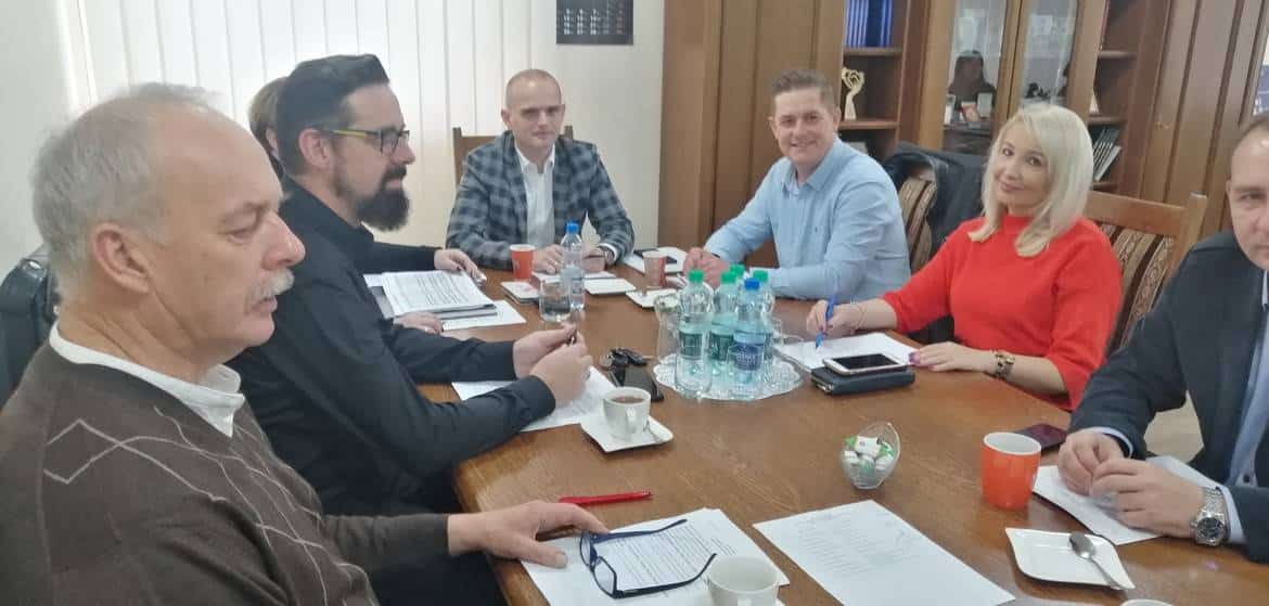 Zdjęcie przedstawia grupę ludzi podczas spotkania biznesowego w biurze, siedzących przy stole z notatnikami i napojami.