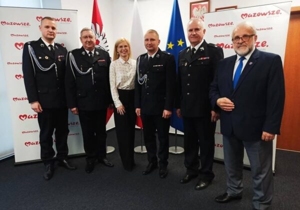 Na zdjęciu znajduje się grupa mężczyzn w mundurach strażackich oraz jedna kobieta, wszyscy pozują na tle flag i banerów z napisem "Mazowsze".