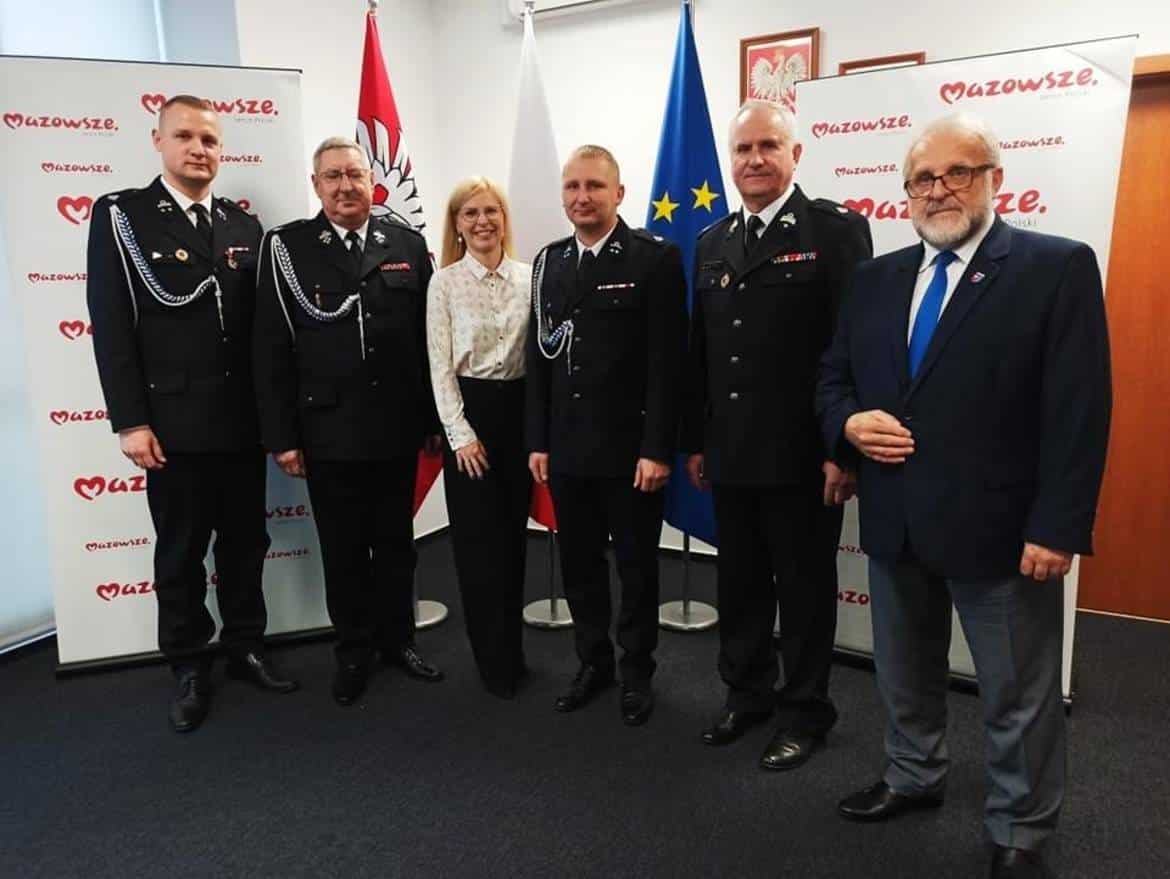 Na zdjęciu znajduje się grupa mężczyzn w mundurach strażackich oraz jedna kobieta, wszyscy pozują na tle flag i banerów z napisem "Mazowsze".