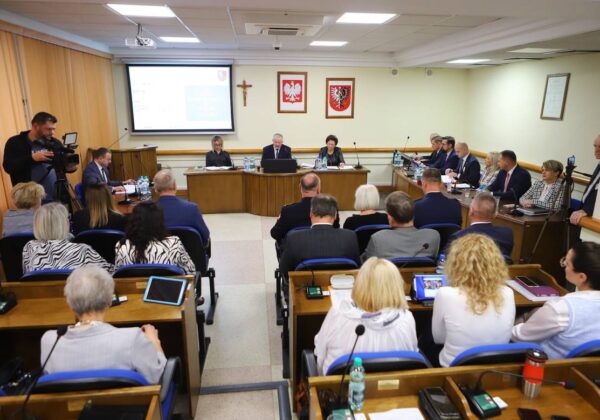 Na zdjęciu przedstawiony jest panel dyskusyjny z uczestnikami siedzącymi w sali obrad, podczas gdy na przodzie prowadzone są prezentacje oraz monitorowane wiadomości.