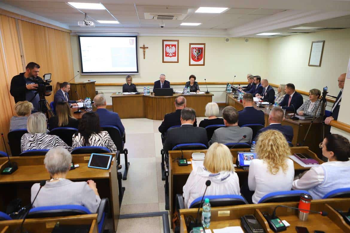 Na zdjęciu przedstawiony jest panel dyskusyjny z uczestnikami siedzącymi w sali obrad, podczas gdy na przodzie prowadzone są prezentacje oraz monitorowane wiadomości.