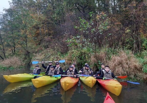 Grupa ludzi w kamizelkach siedzi w kolorowych kajakach na spokojnej rzece w otoczeniu jesiennej przyrody.
