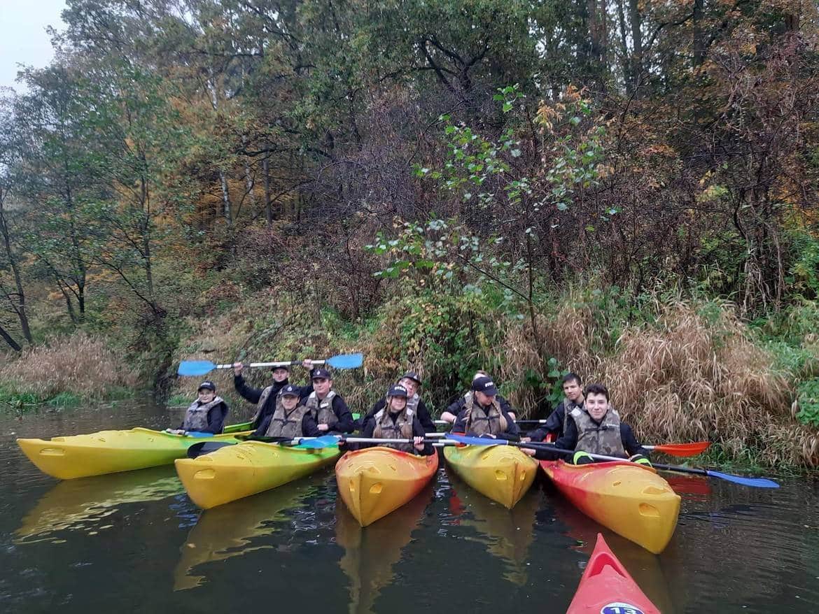 Grupa ludzi w kamizelkach siedzi w kolorowych kajakach na spokojnej rzece w otoczeniu jesiennej przyrody.