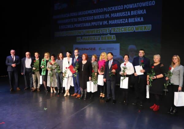 Na zdjęciu znajduje się grupa osób uczestniczących w inauguracji roku akademickiego Płockiego Powiatowego Uniwersytetu Trzeciego Wieku, wszyscy trzymają róże i dokumenty.