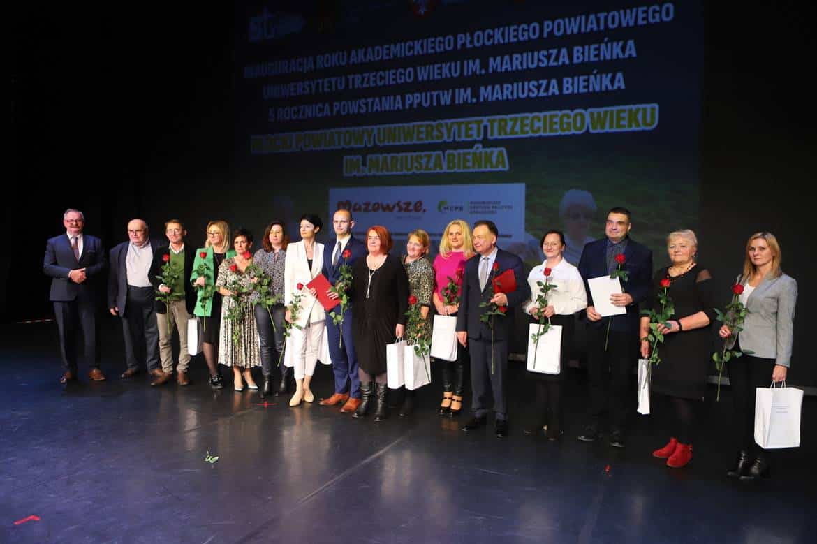 Na zdjęciu znajduje się grupa osób uczestniczących w inauguracji roku akademickiego Płockiego Powiatowego Uniwersytetu Trzeciego Wieku, wszyscy trzymają róże i dokumenty.