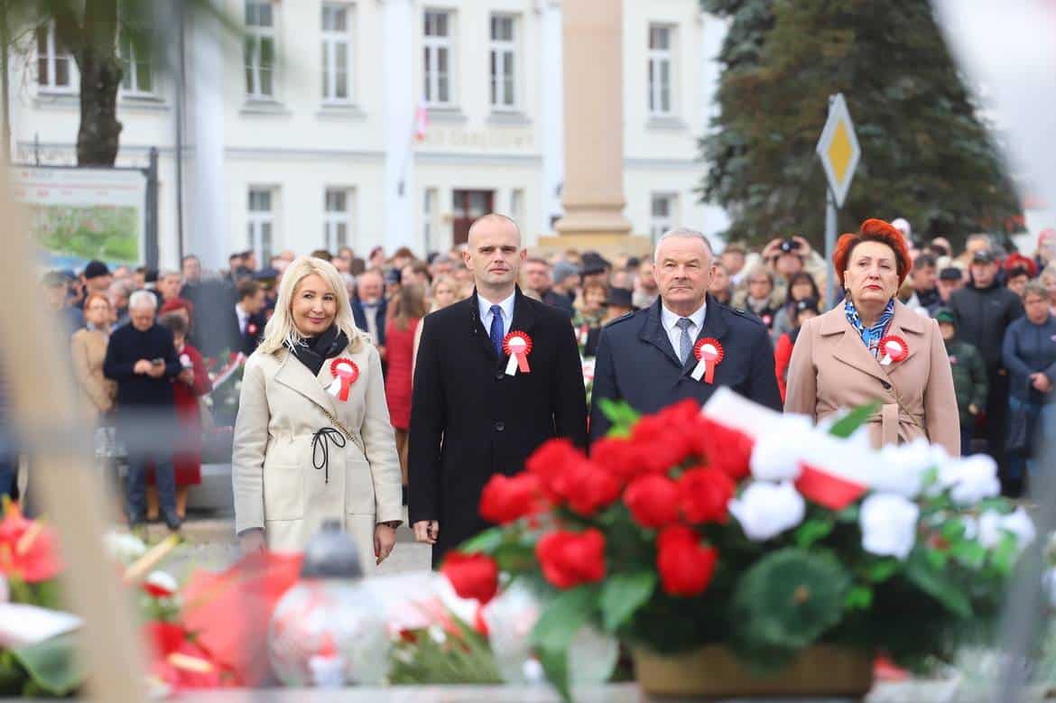 Na zdjęciu widoczna jest grupa osób biorących udział w uroczystości, stojąca przed pomnikiem, z kwiatami i zniczami w pierwszym planie, w tle tłum z flagami i odpowiednimi przypinkami.