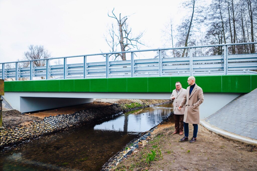 Zdjęcie przedstawia dwóch mężczyzn stojących obok nowego mostu, który łączy drogi nad płynącą rzeką, symbolizując inwestycję oczekiwaną przez mieszkańców od lat.