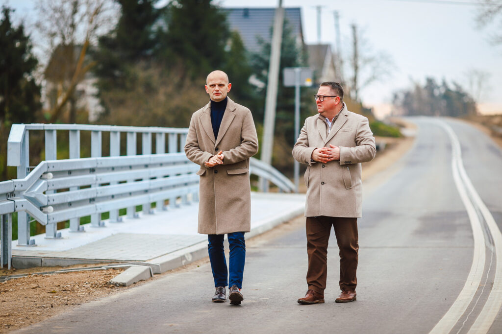 Na zdjęciu przedstawiono dwóch mężczyzn w eleganckich płaszczach spacerujących wzdłuż nowej drogi z dwoma mostami w tle, symbolizujących długo oczekiwaną inwestycję.