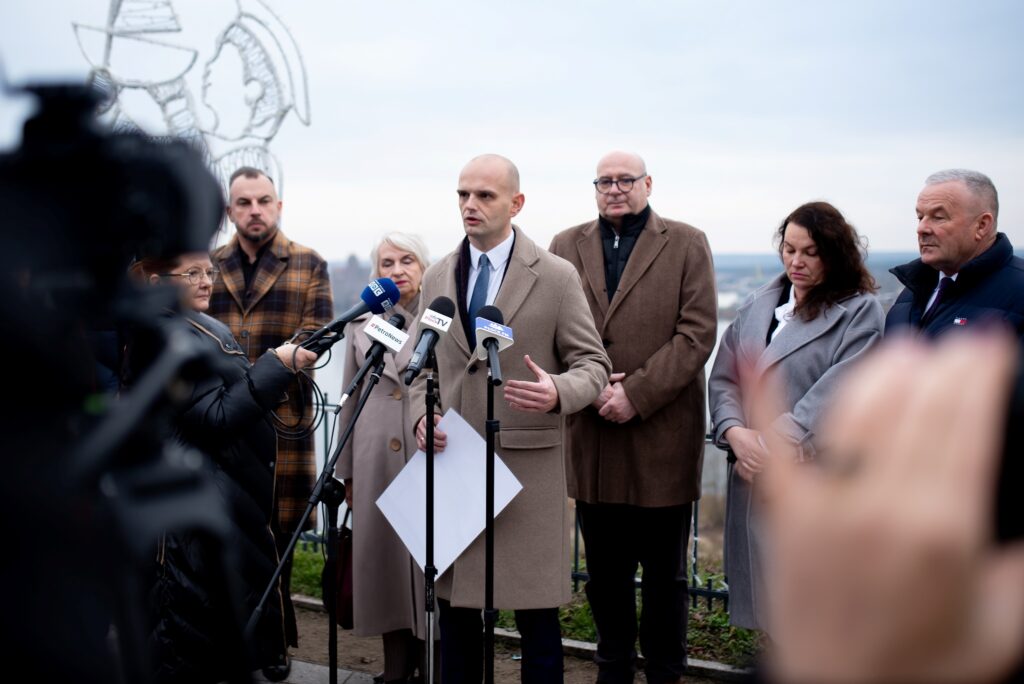 Na zdjęciu widoczny jest starosta Płocki przemawiający podczas konferencji prasowej na temat przywracania bezpieczeństwa powodziowego, otoczony przez zespół współpracowników i dziennikarzy.