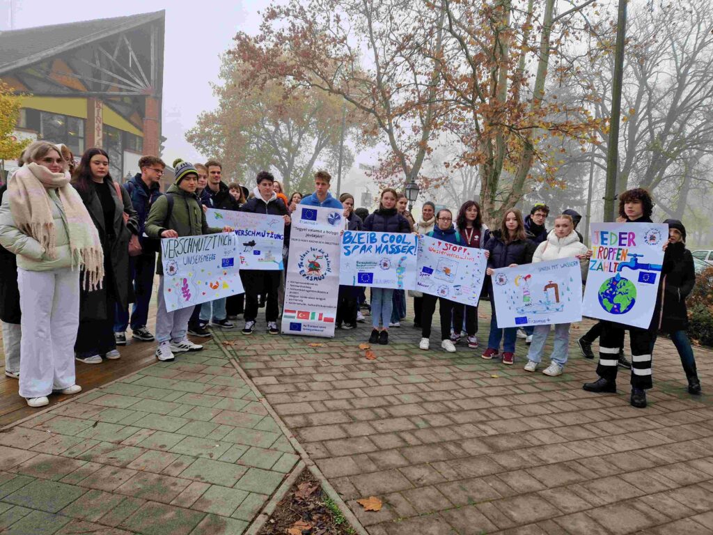 Grupa uczniów z Polski i zagranicy uczestniczy w akcji promującej ochronę wód, trzymając kolorowe transparenty w otoczeniu przyrody na Węgrzech.