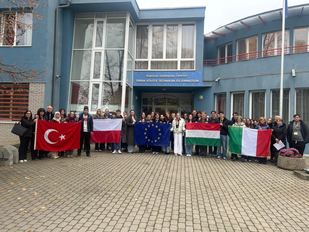 Grupa uczniów i nauczycieli z różnych krajów, trzymających flagi, pozują przed budynkiem szkoły w ramach programu mobilności Erasmus na Węgrzech.