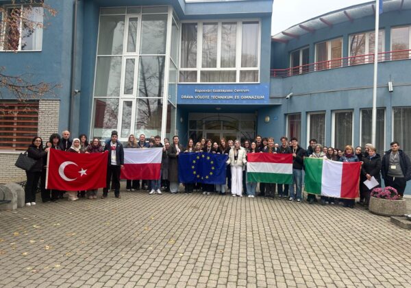 Grupa uczniów i nauczycieli z różnych krajów, trzymających flagi, pozują przed budynkiem szkoły w ramach programu mobilności Erasmus na Węgrzech.