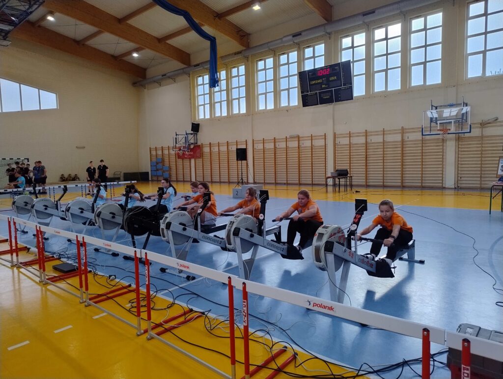 Na zdjęciu widać uczestniczki finału powiatowego zawodów w ergometrze wioślarskim, które rywalizują na urządzeniach w hali sportowej.