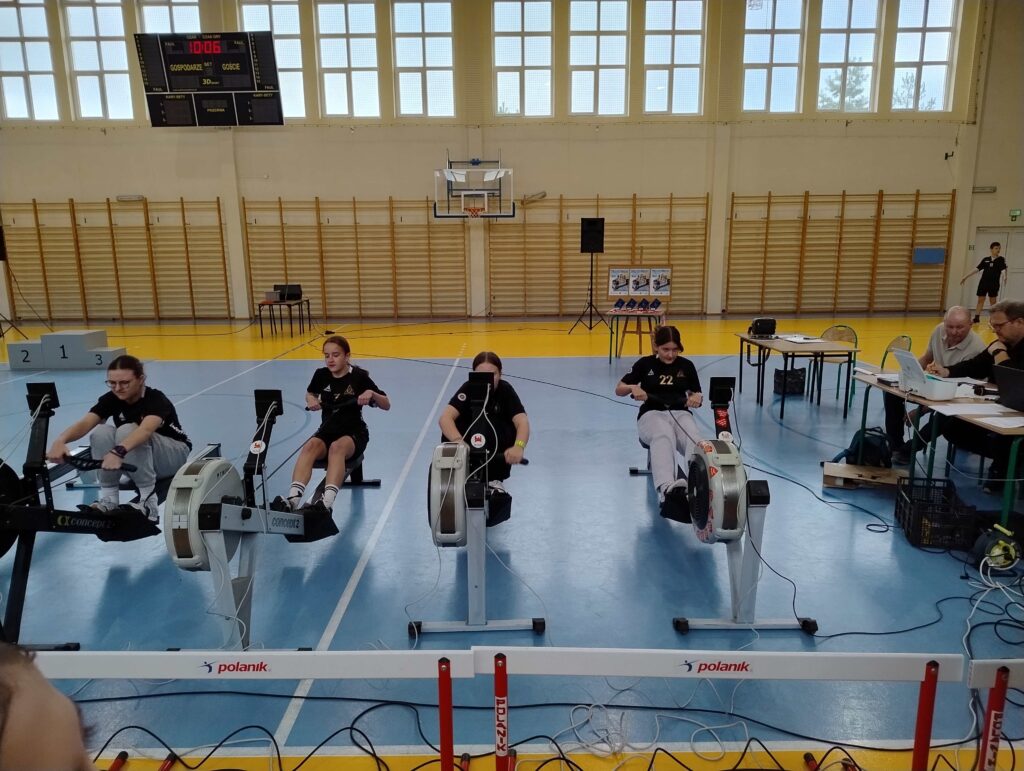 Na zdjęciu przedstawiono uczestników finału powiatowego zawodów w ergometrze wioślarskim, którzy intensywnie rywalizują na urządzeniach w hali sportowej.