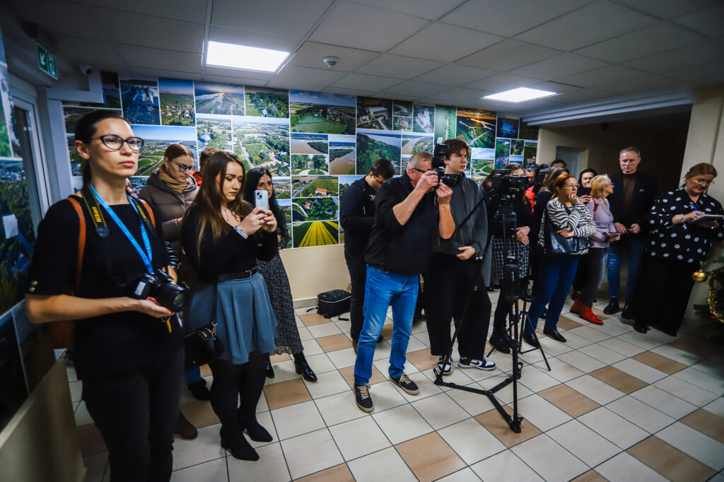 Na zdjęciu widać grupę osób z aparatami i telefonami, zgromadzonych w pomieszczeniu z fotografiami krajobrazów, podczas podpisywania aktu notarialnego dotyczącego wojska w powiecie płockim.