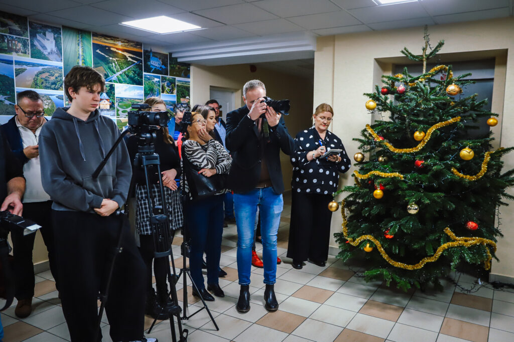 W sali, w której odbywa się wydarzenie związane z podpisaniem aktu notarialnego, zgromadziła się grupa osób, w tym dziennikarze i uczestnicy, a w tle widać udekorowaną choinkę.