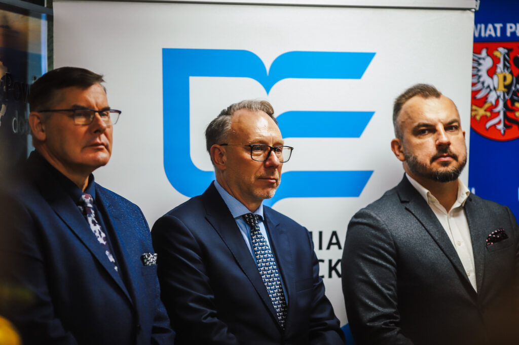 Trzech mężczyzn w garniturach stoi obok siebie przed tłem z logo, wyrażając powagę chwili związanej z podpisaniem aktu notarialnego dotyczącego wojska w powiecie płockim.