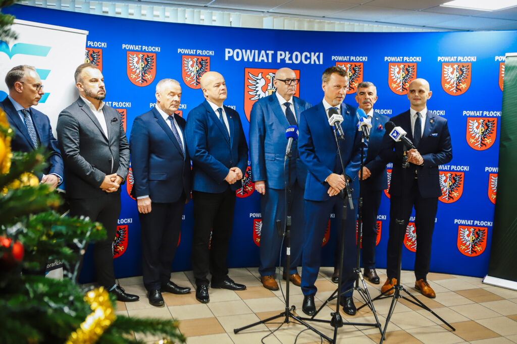 Na zdjęciu widoczna jest grupa mężczyzn ubranych w garnitury, stojących przed niebieskim tłem z herbem powiatu płockiego, podczas wydarzenia związanego z podpisaniem aktu notarialnego dotyczącego wojska w powiecie płockim.