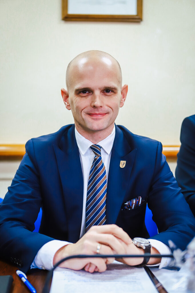 Na zdjęciu widać mężczyznę w granatowym garniturze, który uśmiecha się, siedząc przy stole w trakcie sesji budżetowej Radnych Powiatu Płockiego.