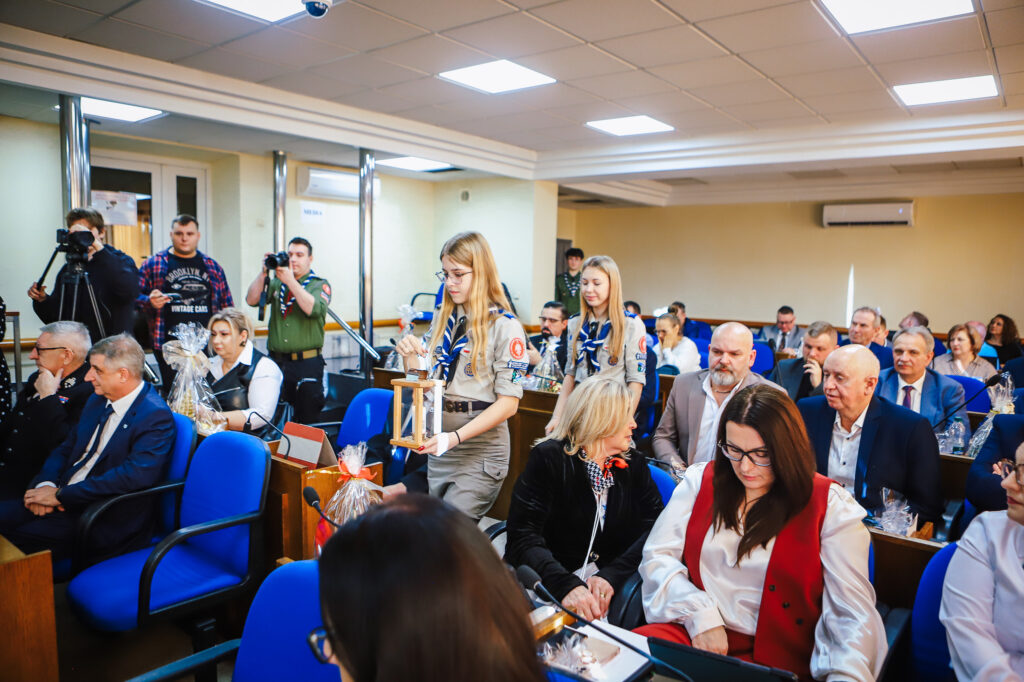 Na zdjęciu widać radnych Powiatu Płockiego zgromadzonych podczas sesji, gdzie uchwalają rekordowy budżet, a dwie harcerki wchodzą z lampionem.