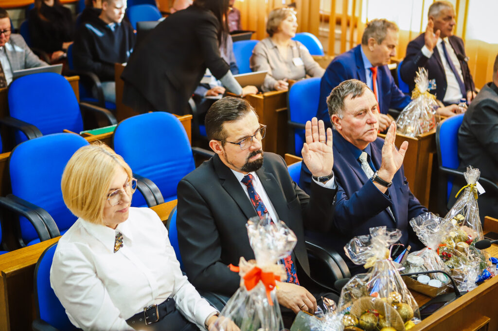 Radni Powiatu Płockiego podczas sesji, na której uchwalono rekordowy budżet, siedzą w sali obrad z uniesionymi rękami.