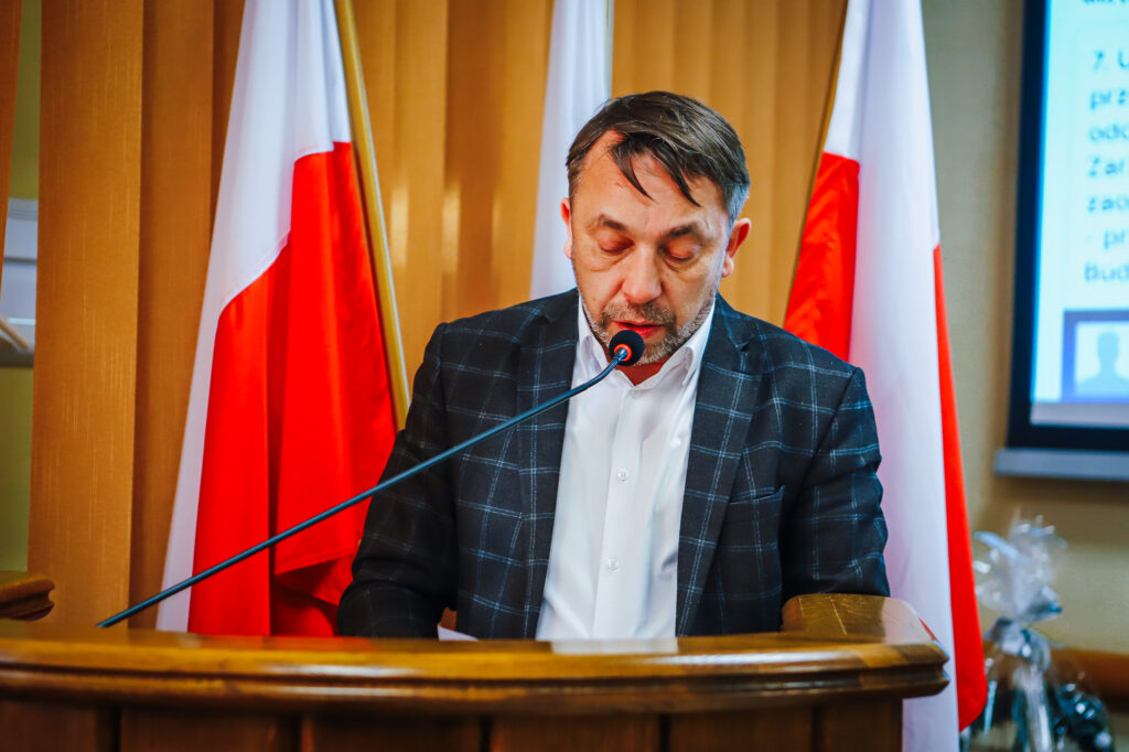 Radny Powiatu Płockiego podczas sesji odczytuje najważniejsze informacje dotyczące uchwały o rekordowym budżecie, z flagami polskimi w tle.