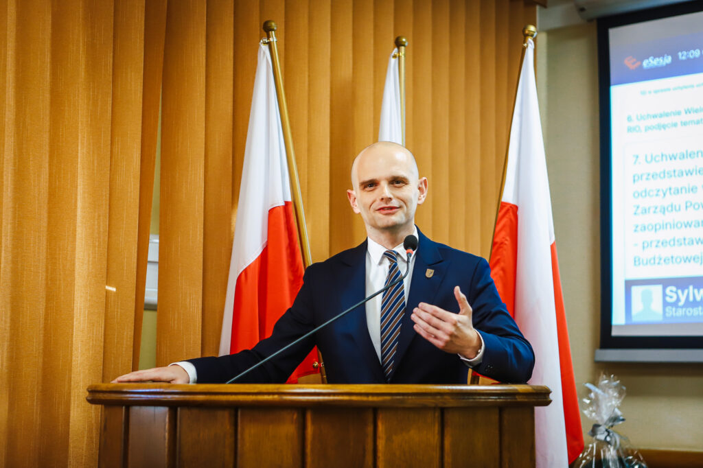Na zdjęciu przedstawiony jest mężczyzna w garniturze, przemawiający z mównicy, obok której znajdują się flagi, podczas obrad Radnych Powiatu Płockiego dotyczących uchwały budżetowej.