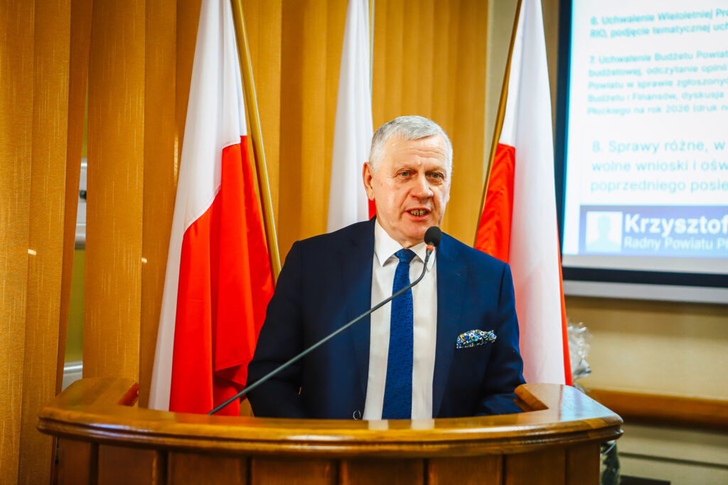 Na zdjęciu wystąpienie radnego Powiatu Płockiego, który ogłasza uchwałę o rekordowym budżecie, z flagami Polski w tle.