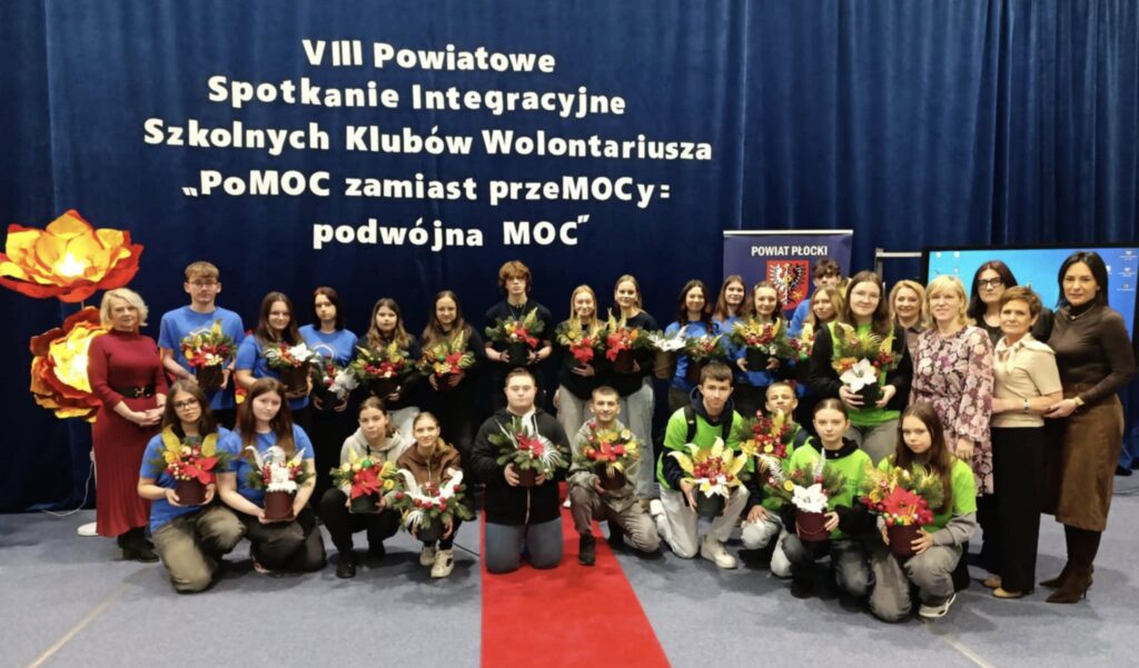 Uczestnicy VIII Powiatowego Spotkania Integracyjnego Szkolnych Klubów Wolontariusza trzymają kwiaty na tle niebieskiego banera z hasłem wydarzenia.