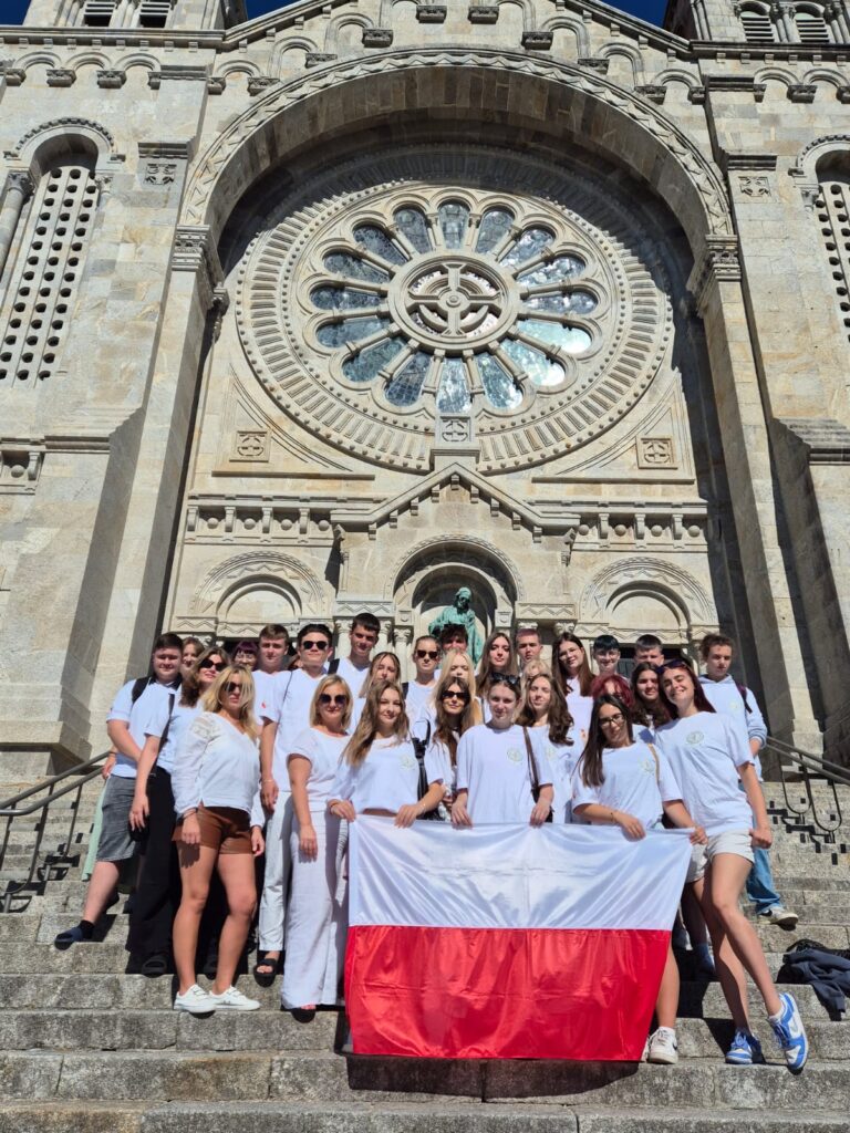 Uczestnicy projektu „Dobre praktyki zagraniczne jako klucz do sukcesu” pozują na schodach przed imponującą budowlą, trzymając flagę Polski.