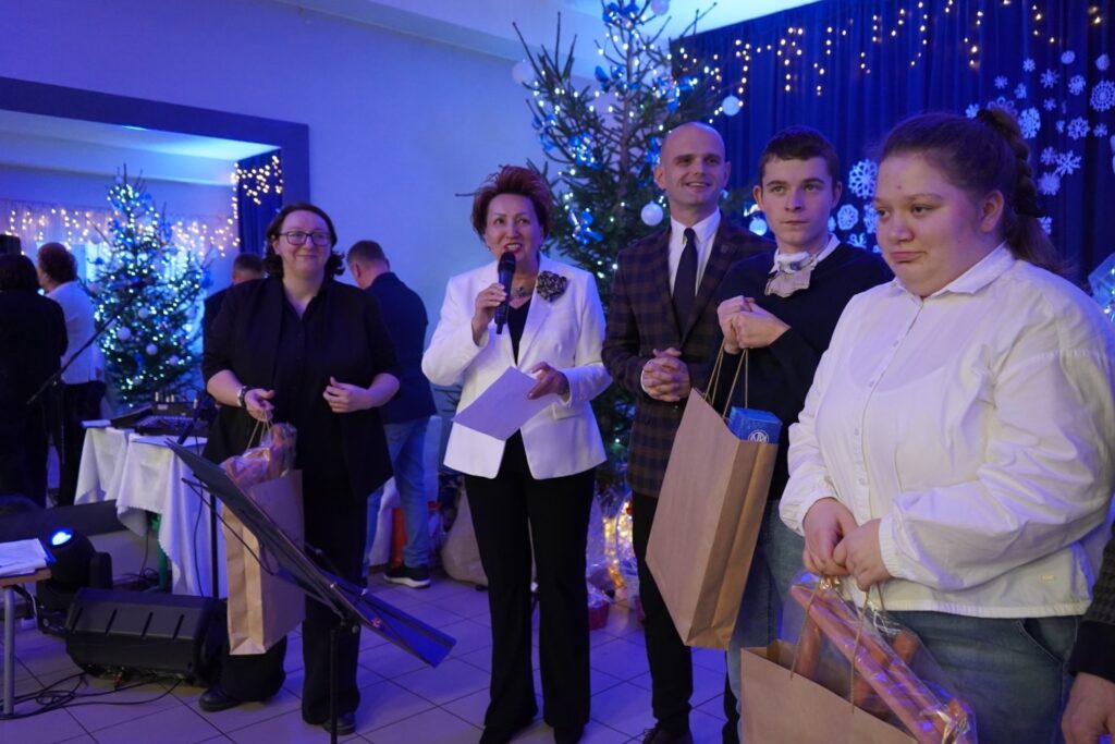 Na zdjęciu nowa Pani Starosta przemawia do zgromadzonych, trzymając kartki, a obok niej stoją osoby z upominkami w rękach w świątecznej scenerii.