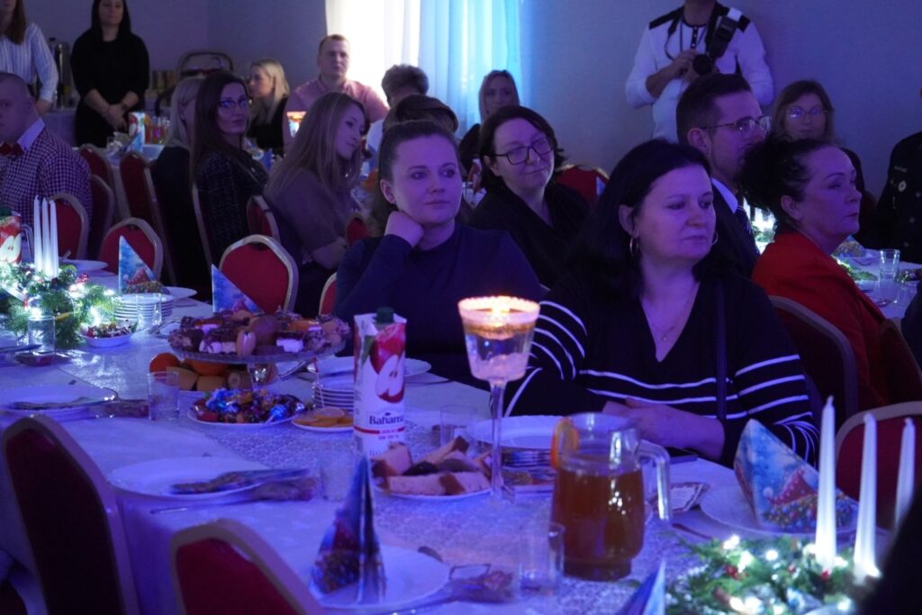 Zdjęcie przedstawia grupę gości podczas uroczystości, w której uczestniczą w elegancko udekorowanej sali, skupieni na wydarzeniu.