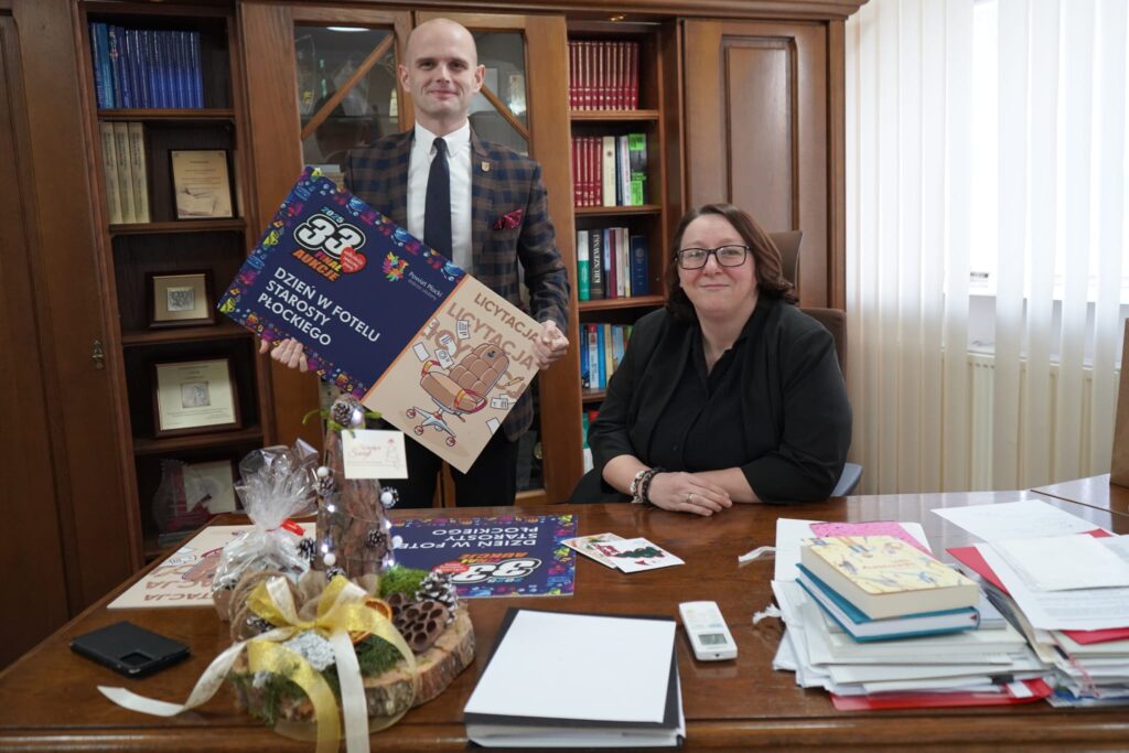 Na zdjęciu nowa Pani Starosta prezentuje plakaty związane z inicjatywą "Dzień w fotelu Starosty Płockiego" oraz "Licytacja", siedząc przy biurku w swoim biurze.