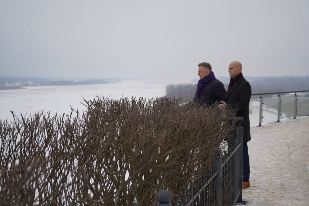 Wiceminister i przedstawiciel samorządu stoją przy barierce, podziwiając zimowy krajobraz nad rzeką w Powiecie Płockim.
