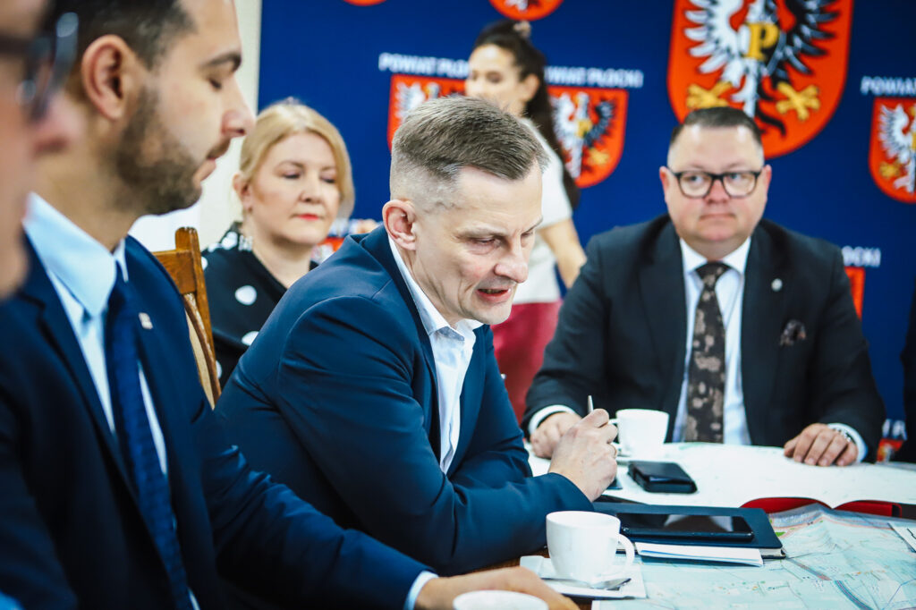 Na zdjęciu przedstawiono spotkanie grupy osób dyskutujących nad projektami związanymi z trasą S10, w otoczeniu map i filiżanek kawy.