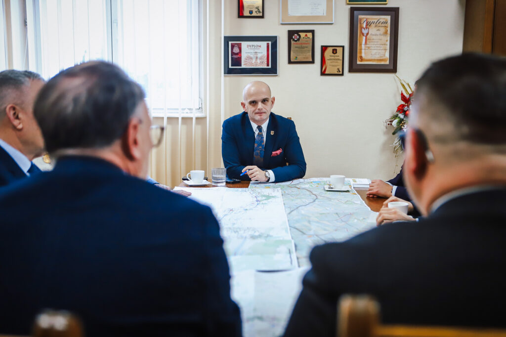 W spotkaniu poświęconym trasie S10, z udziałem mężczyzn w garniturach, dominujący jest mężczyzna w jasnym garniturze, siedzący przy stole z mapami i filiżankami.