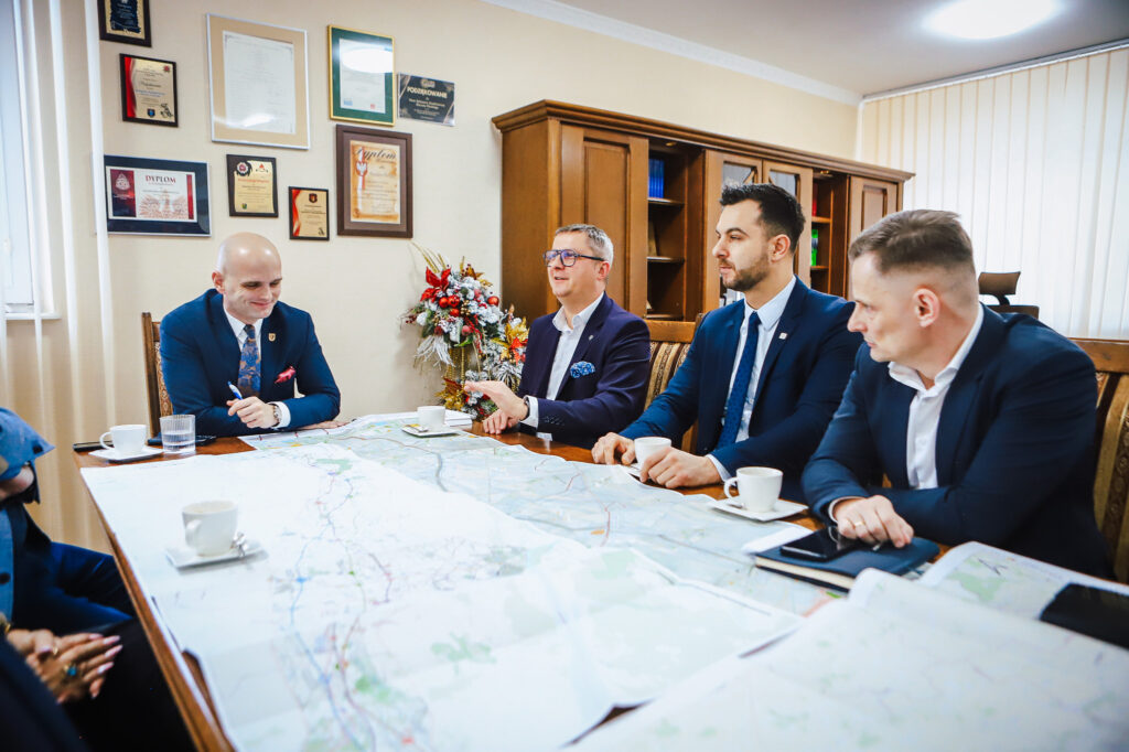 Na zdjęciu przedstawiono spotkanie czterech mężczyzn przy stole wypełnionym mapami, w biurze udekorowanym dyplomami i kwiatami.