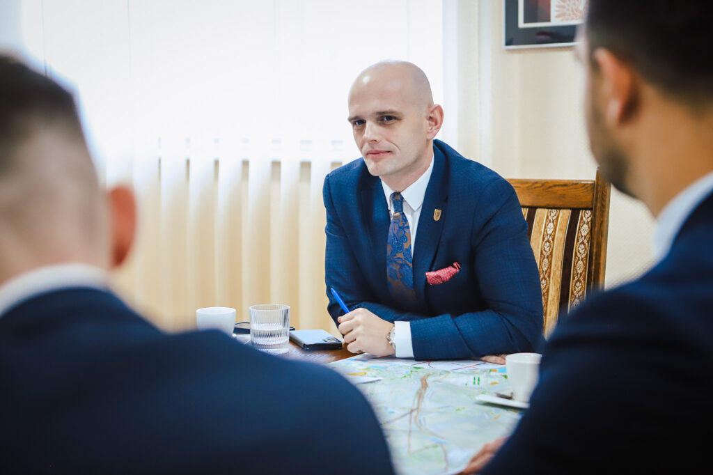 Na zdjęciu widoczna jest mężczyzna w garniturze, siedzący przy stole, otoczony innymi uczestnikami spotkania, podczas omawiania planów dotyczących trasy S10 w pobliżu Płocka.