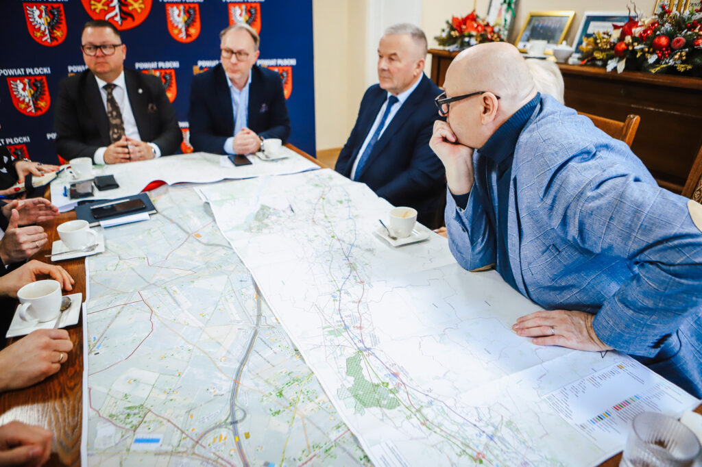 Na zdjęciu przedstawiono spotkanie grupy mężczyzn przy stole, na którym rozłożona jest mapa, a w tle widoczne są elementy dekoracyjne i herb powiatu płockiego.