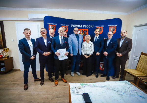 Na zdjęciu widoczna jest grupa dziesięciu osób stojących w sali z logo Powiatu Płockiego, które zapowiadają szczegóły dotyczące trasy S10, niedaleko Płocka.