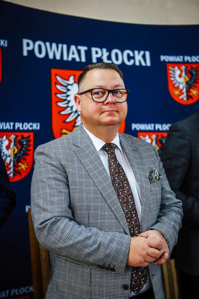 Na zdjęciu widoczny jest mężczyzna w eleganckim garniturze, stojący w pobliżu logotypu Powiatu Płockiego.