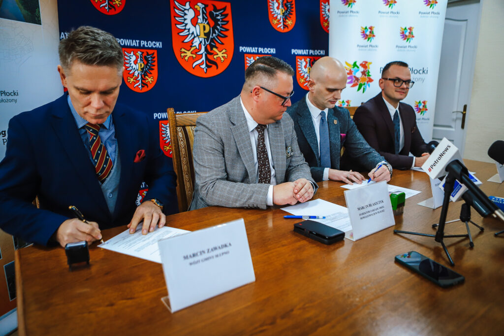 Na zdjęciu widoczna jest ceremonia podpisania umowy, w której biorą udział cztery osoby siedzące przy stole, symbolizująca powstanie nowej ścieżki rowerowej w Powiecie Płockim.