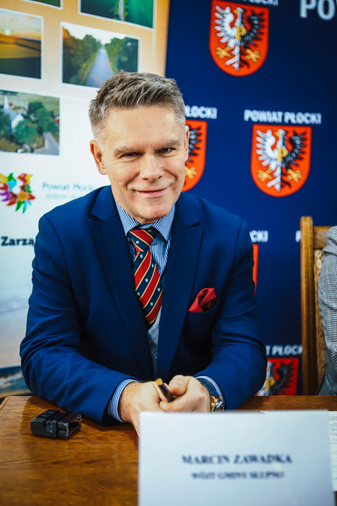 Na zdjęciu widoczny jest mężczyzna w garniturze, uśmiechający się podczas konferencji w Powiecie Płockim, na tle logo powiatu i zdjęć przedstawiających krajobrazy.