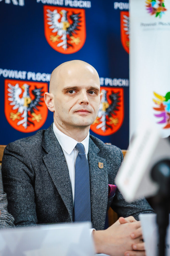 Na zdjęciu widać mężczyznę siedzącego przy stole w kontekście ogłoszenia o nowej ścieżce rowerowej w Powiecie Płockim, z herbem powiatu w tle.