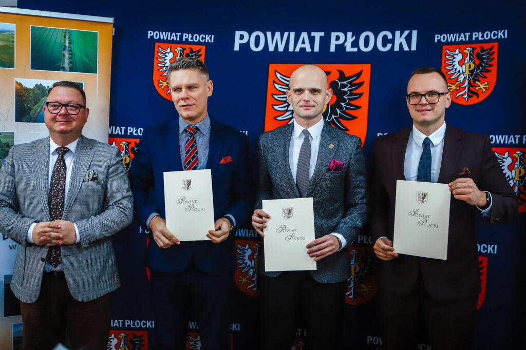 Na zdjęciu czterech mężczyzn w garniturach stoi obok siebie, trzymając dokumenty, z tłem przedstawiającym logotyp Powiatu Płockiego.