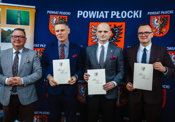 Na zdjęciu czterech mężczyzn w garniturach stoi obok siebie, trzymając dokumenty, z tłem przedstawiającym logotyp Powiatu Płockiego.