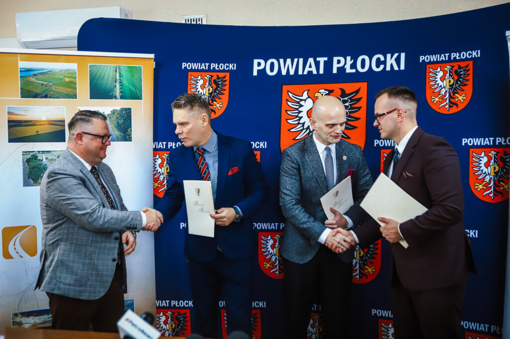 Uroczystość podpisania dokumentów dotyczących budowy nowej ścieżki rowerowej w Powiecie Płockim, z uczestnikami uściskiem dłoni.